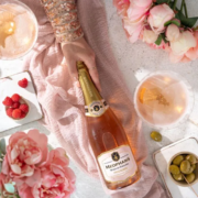 Bottle of Meophams Phoenix sparkling wine on a pink blanket, surrounded by raspberries, pink peonies, candles, and a glass of wine.
