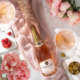 Bottle of Meophams Phoenix sparkling wine on a pink blanket, surrounded by raspberries, pink peonies, candles, and a glass of wine.