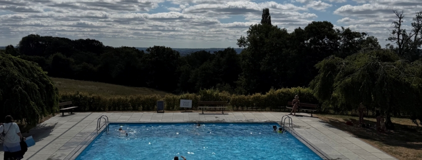 Outdoor swimming pool with bright blue water and clear sky, surrounded by trees and a paved deck with a few people.